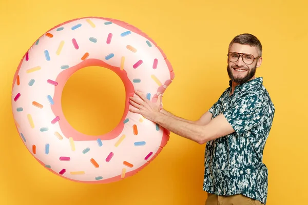 Side view of smiling bearded guy in glasses with donut swim ring on yellow — Stock Photo