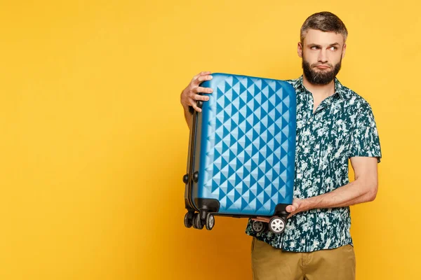 Грустный бородатый парень с голубым чемоданом на желтом — стоковое фото