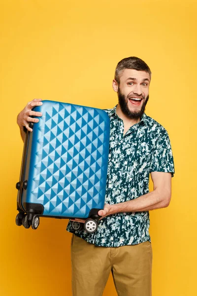 Happy bearded guy with blue suitcase on yellow — Stock Photo