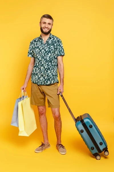 Повний вигляд щасливого бородатого хлопця з валізою і сумками для покупок на жовтому — стокове фото