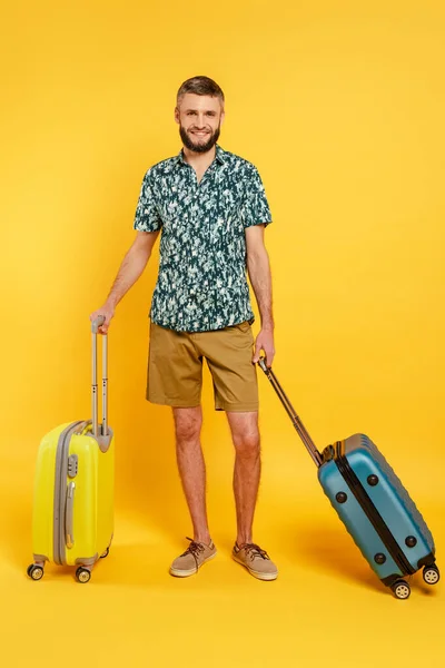 Повний вигляд щасливого бородатого хлопця з дорожніми сумками на жовтому — стокове фото
