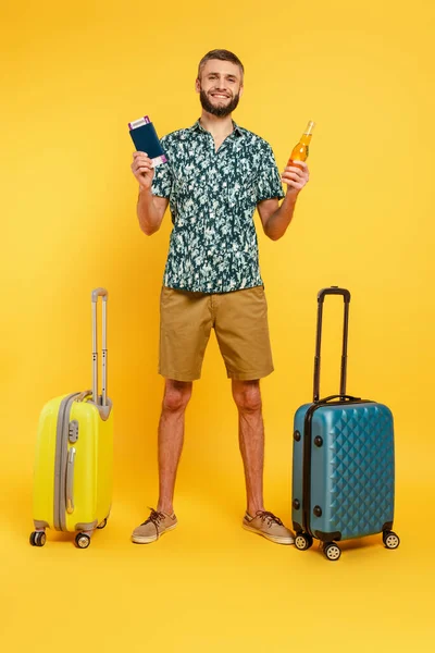 Вид в полный рост счастливого бородатого парня с дорожными сумками, пивом и паспортом на желтом — стоковое фото