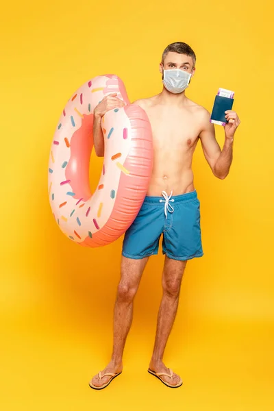 Vue pleine longueur du gars dans les maillots de bain et masque médical avec anneau de natation et passeport sur jaune — Photo de stock