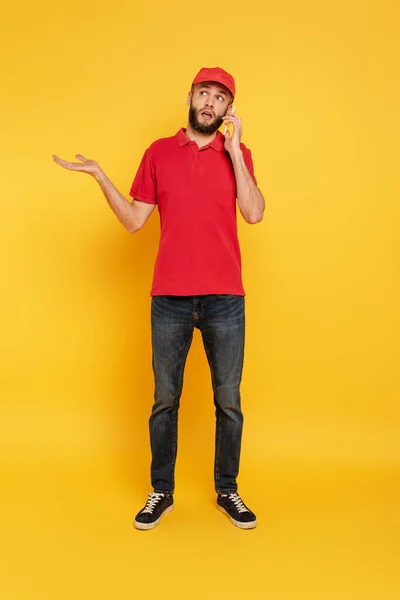 Confundido barbudo repartidor hombre en uniforme rojo hablando en el teléfono inteligente en amarillo - foto de stock