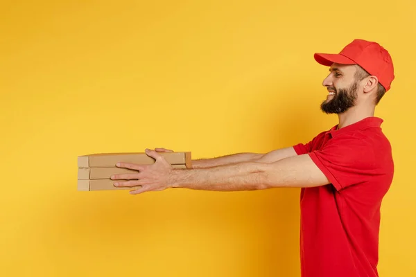 Vista lateral del repartidor barbudo sonriente hombre en uniforme rojo dando cajas de pizza en amarillo - foto de stock