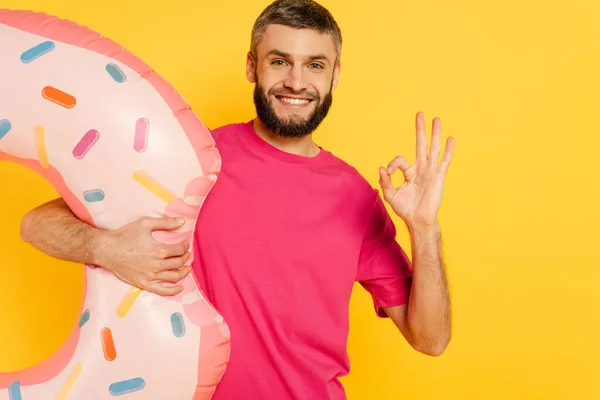 Happy bearded guy in pink t-shirt with swim ring showing ok sign isolated on yellow — Stock Photo