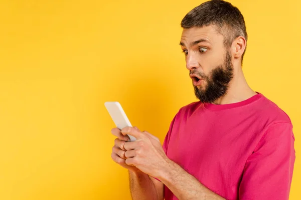 Cara barbudo chocado em camiseta rosa usando smartphone no amarelo — Fotografia de Stock