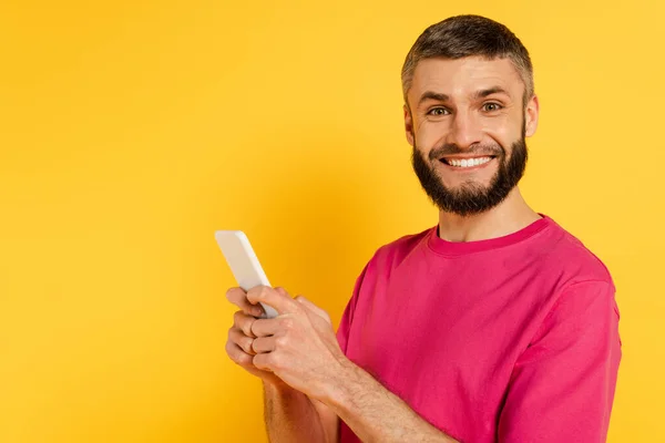 Glücklicher bärtiger Kerl im rosa T-Shirt mit Smartphone auf gelb — Stockfoto