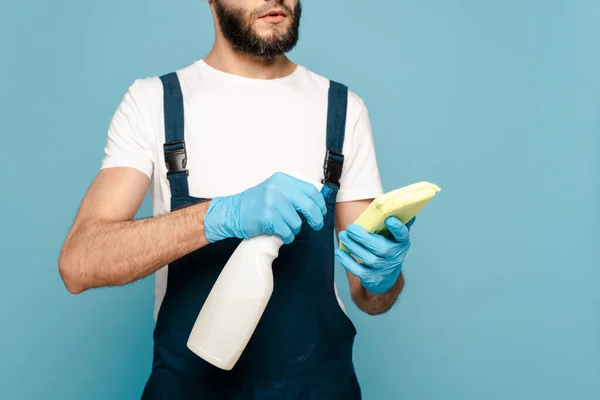 Vista recortada del limpiador en uniforme y guantes de goma con detergente y esponja sobre fondo azul - foto de stock