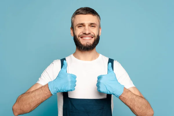 Щасливий прибиральник в уніформі і гумових рукавичках, що показують великі пальці вгору на синьому фоні — стокове фото