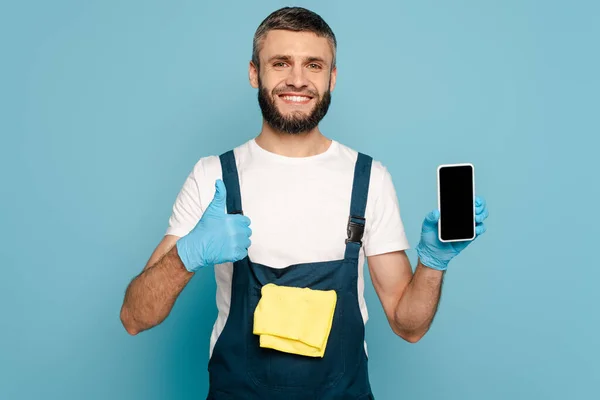 Limpiador feliz en uniforme y guantes de goma con alfombra que sostiene el teléfono inteligente y muestra el pulgar hacia arriba sobre fondo azul - foto de stock