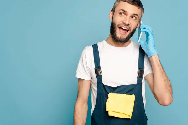 Excited cleaner in uniform and rubber gloves with rug talking on smartphone isolated on blue — Stock Photo