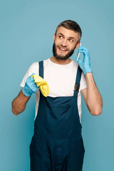 Felice detergente in uniforme e guanti di gomma con tappeto parlare su smartphone isolato su blu — Foto stock