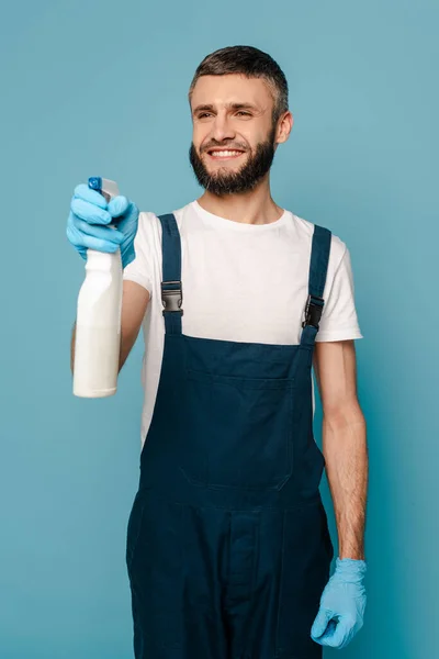 Limpador feliz em uniforme e luvas de borracha segurando detergente spray no fundo azul — Fotografia de Stock