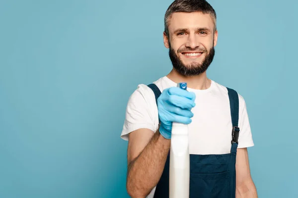 Limpador feliz em uniforme e luvas de borracha segurando detergente spray no fundo azul — Fotografia de Stock