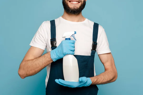 Обрезанный вид счастливого очистителя в форме и резиновых перчатках держа спрей моющее средство на синем фоне — стоковое фото