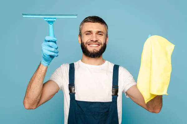 Limpiador feliz en uniforme y guantes de goma sujetando trapo y escobilla sobre fondo azul - foto de stock