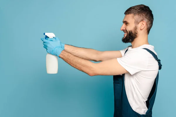 Visão lateral do limpador feliz em uniforme e luvas de borracha segurando detergente spray no fundo azul — Fotografia de Stock
