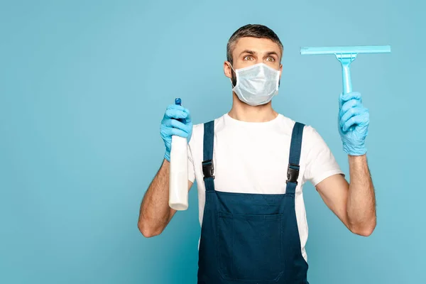 Reiniger in medizinischer Maske mit Reinigungsmittel und Rakel auf blauem Hintergrund — Stock Photo