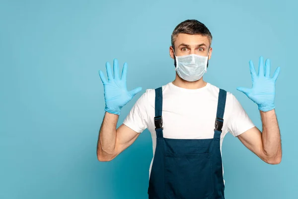 Reinigungskraft in medizinischer Maske zeigt Hände in Gummihandschuhen auf blauem Hintergrund — Stockfoto