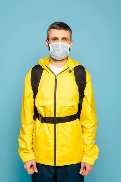 Repartidor en máscara médica y uniforme amarillo con mochila sobre fondo azul - foto de stock