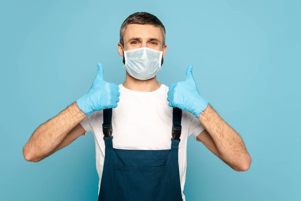 Limpador em máscara médica mostrando polegares para cima no fundo azul — Fotografia de Stock