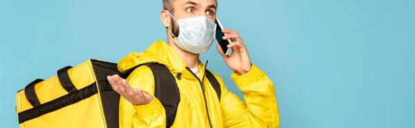 Repartidor confuso en máscara médica y uniforme amarillo con mochila hablando en teléfono inteligente aislado en azul - foto de stock