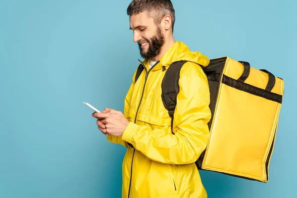Repartidor sonriente en uniforme amarillo con mochila usando teléfono inteligente sobre fondo azul - foto de stock