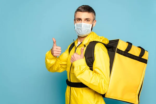 Repartidor en máscara médica y uniforme amarillo con mochila mostrando el pulgar hacia arriba sobre fondo azul - foto de stock