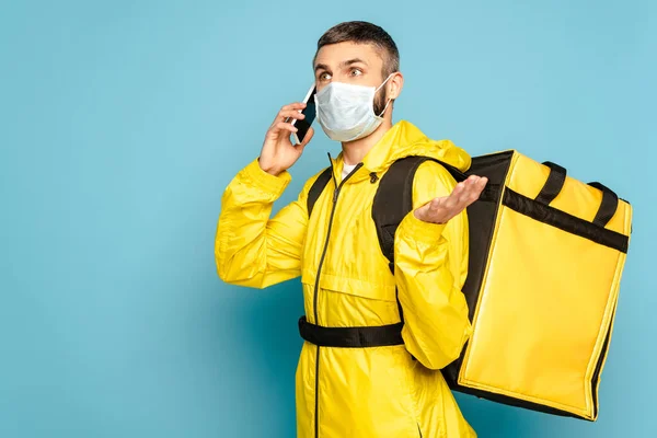 Смущенный курьер в медицинской маске и желтой форме с рюкзаком разговаривает на смартфоне на синем фоне — стоковое фото