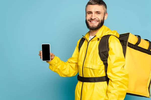 Entregador feliz em uniforme amarelo com mochila mostrando smartphone com tela em branco no fundo azul — Fotografia de Stock