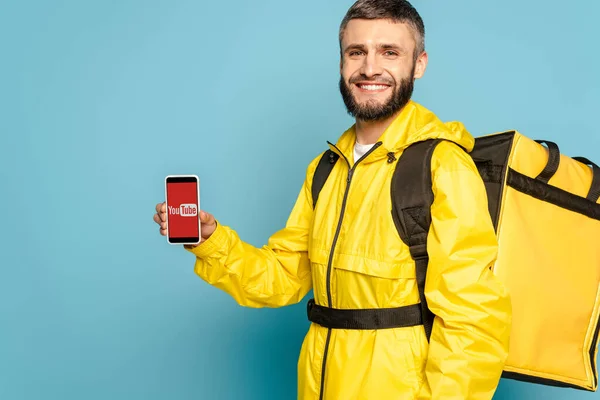 KYIV, UCRANIA - 30 DE MARZO DE 2020: repartidor feliz en uniforme amarillo con mochila que muestra el teléfono inteligente con aplicación youtube sobre fondo azul - foto de stock