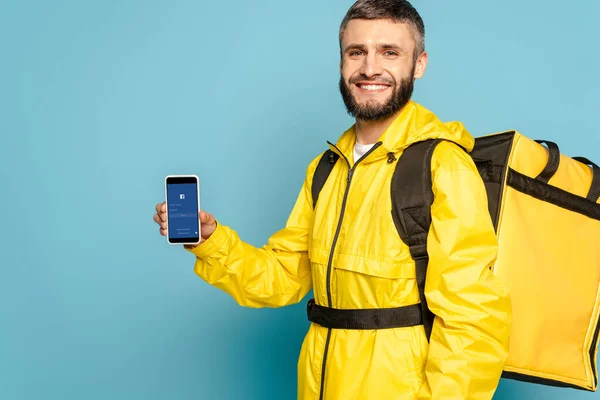 KYIV, UCRANIA - 30 DE MARZO DE 2020: repartidor feliz en uniforme amarillo con mochila que muestra el teléfono inteligente con aplicación de facebook sobre fondo azul - foto de stock