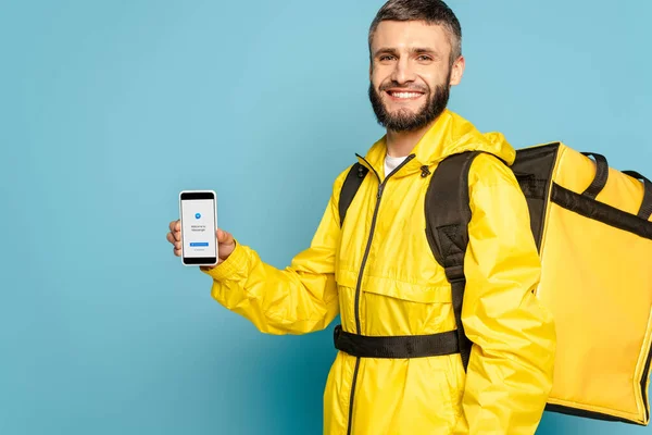 KYIV, UKRAINE - 30 MARS 2020 : livreur heureux en uniforme jaune avec sac à dos montrant smartphone avec application messager sur fond bleu — Photo de stock