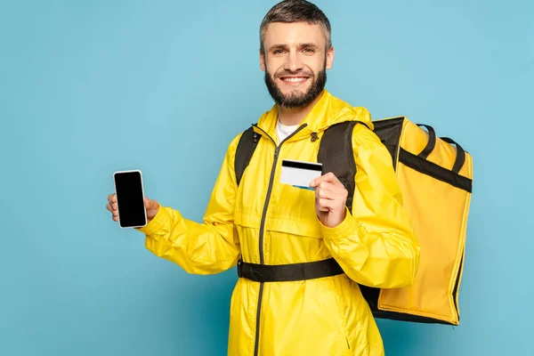 Happy deliveryman in yellow uniform with backpack showing smartphone with blank screen while holding credit card on blue background — Stock Photo