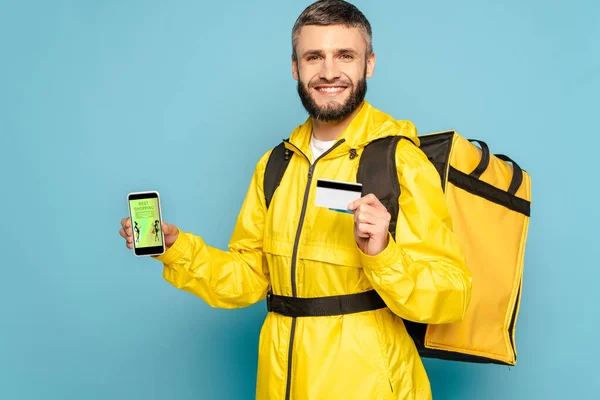 Щасливий рятівник в жовтій формі з рюкзаком, що показує смартфон із додатком для покупок моди, тримаючи кредитну картку на синьому фоні — стокове фото