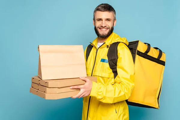 Lächelnder Auslieferer in gelber Uniform mit Rucksack mit Papierverpackung und Pizzakartons auf blauem Hintergrund — Stockfoto