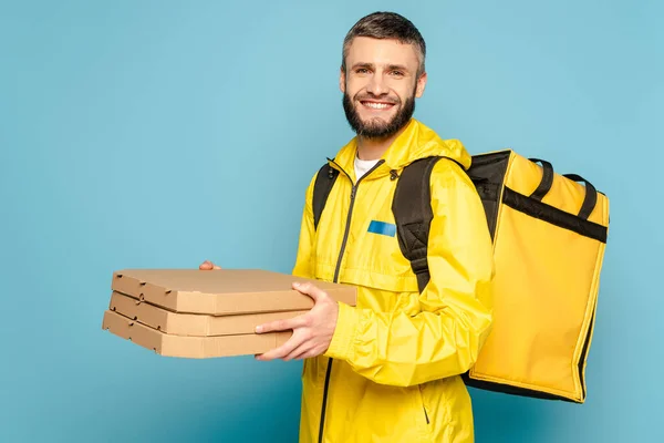 Livreur souriant en uniforme jaune avec sac à dos tenant des boîtes à pizza sur fond bleu — Photo de stock