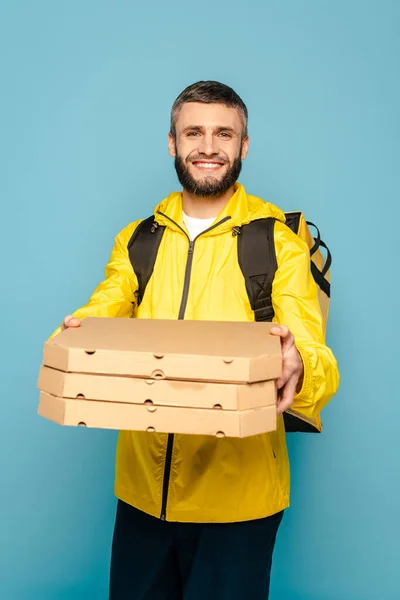 Fattorino sorridente in uniforme gialla con zaino contenente scatole di pizza su sfondo blu — Foto stock