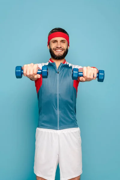 Усміхнений стильний спортсмен займається з гантелями на синьому фоні — стокове фото