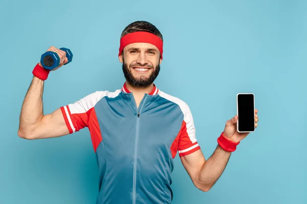 Sonriente deportista con estilo haciendo ejercicio con mancuerna y sosteniendo el teléfono inteligente sobre fondo azul — Stock Photo