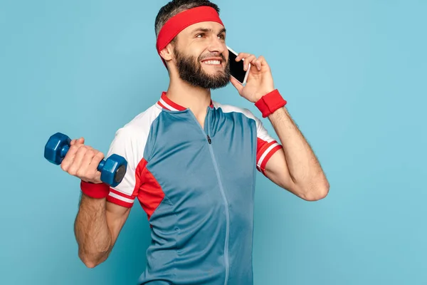 Smiling stylish sportsman exercising with dumbbell and talking on smartphone on blue background — Stock Photo