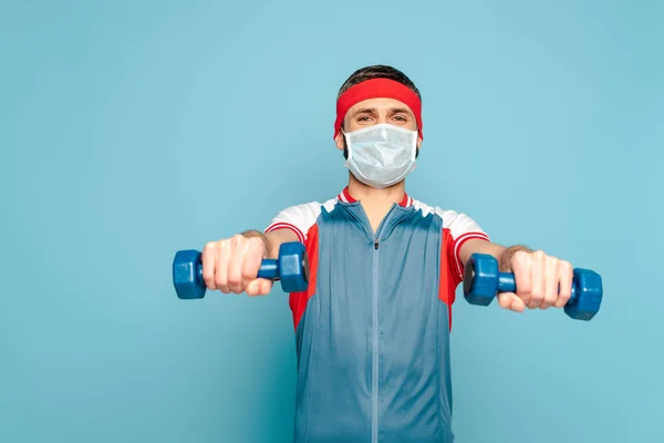 Sportif élégant dans le masque médical exercice avec haltères sur fond bleu — Photo de stock