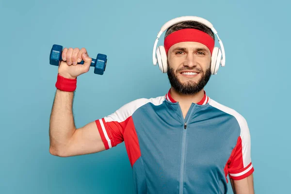 Щасливий стильний спортсмен в навушниках вправляється з гантелі на синьому фоні — стокове фото
