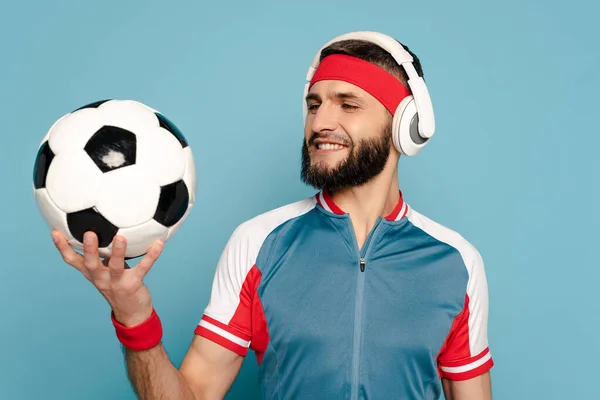 Deportista con estilo feliz en auriculares con pelota de fútbol sobre fondo azul - foto de stock