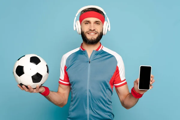 Deportista con estilo feliz en auriculares con pelota de fútbol y teléfono inteligente sobre fondo azul - foto de stock