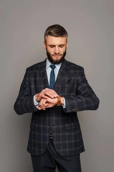 Schöner bärtiger Geschäftsmann im Anzug mit Blick auf die Armbanduhr auf grauem Hintergrund — Stockfoto