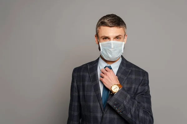 Guapo hombre de negocios en máscara médica ajustando corbata sobre fondo gris - foto de stock