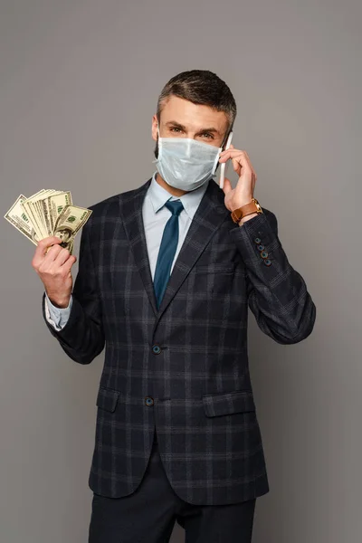 Hombre de negocios guapo en máscara médica con dinero hablando en el teléfono inteligente sobre fondo gris - foto de stock