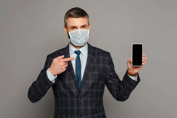 Handsome businessman in medical mask pointing with finger at smartphone with blank screen on grey background — Stock Photo